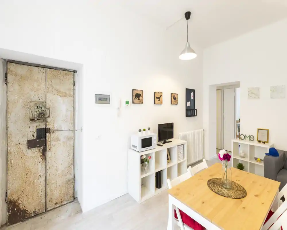 Bright, modern living/dining area with a wooden dining table, shelving unit with microwave and TV, and tasteful decorative accents providing a welcoming atmosphere.