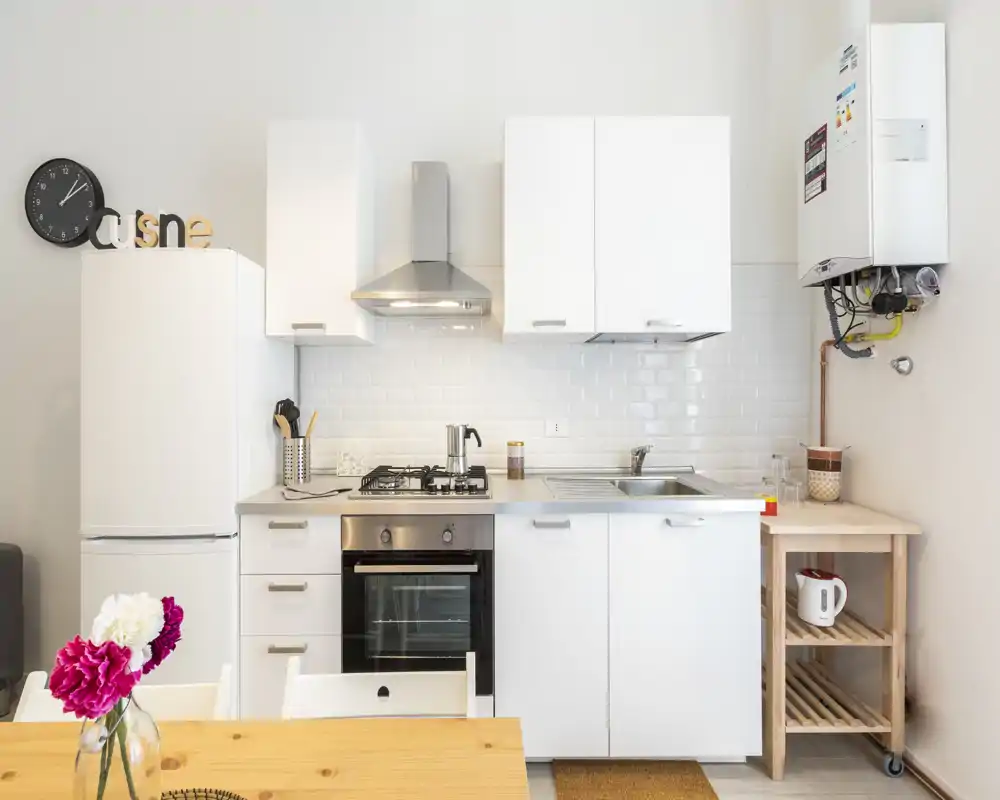 Bright, modern kitchenette with white cabinets, built-in oven and gas hob, stainless-steel sink and tasteful wooden table with fresh flowers.