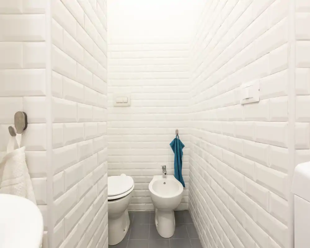 Compact, modern bathroom with white subway-tiled walls, a toilet and a bidet, and a turquoise hand towel adding a touch of color.