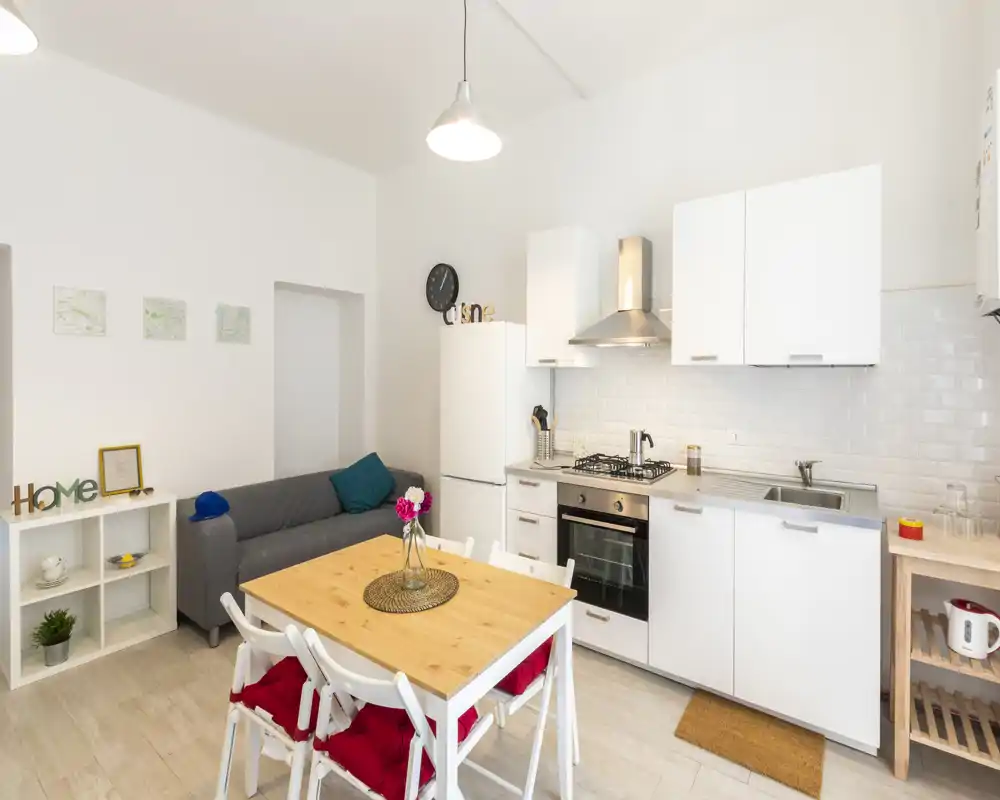 Bright, modern open-plan kitchen with white cabinets, gas hob, oven and a wooden dining table set; a small sofa and decorative cushions create a cozy studio layout.