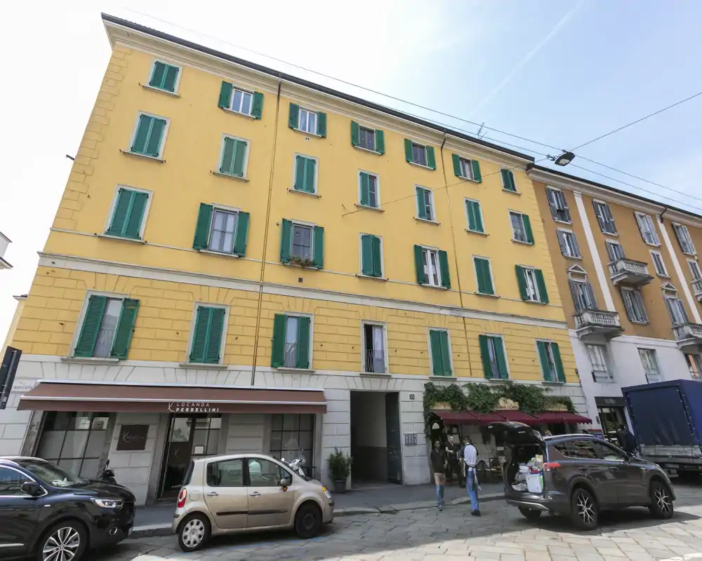 Street-level exterior of a yellow apartment building with green shutters, ground-floor shops and parked cars — useful for showing the building facade and neighborhood context.