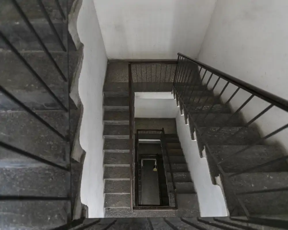 A top-down view of a multi-storey stairwell with stone steps and metal railings, showing the building's internal staircase and landings.