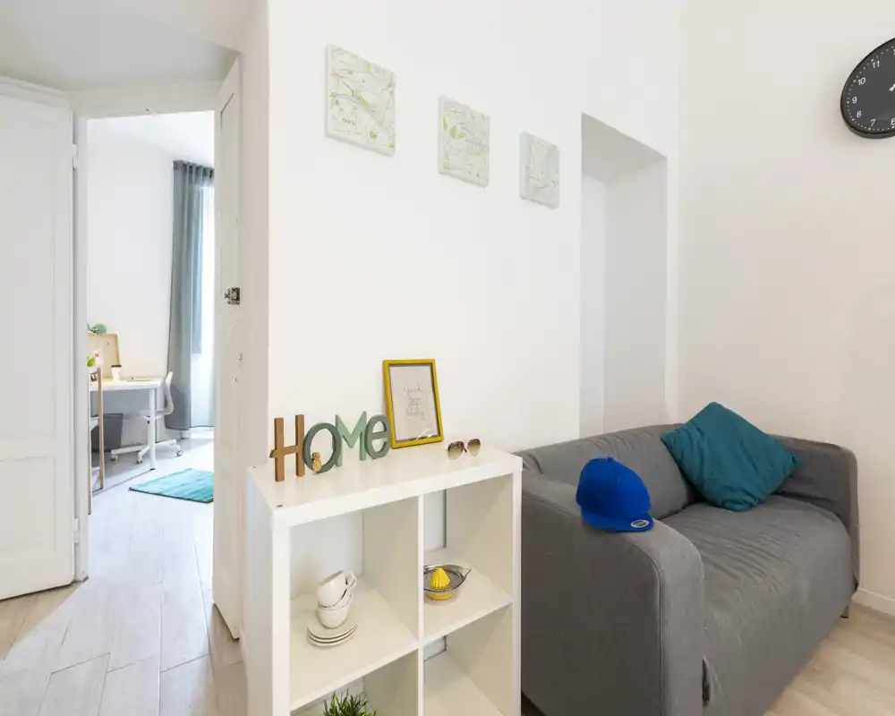 Bright, modern living area with a compact grey sofa, teal cushion and decorative shelving; the photo shows part of an adjacent room with natural light, creating an airy feel.