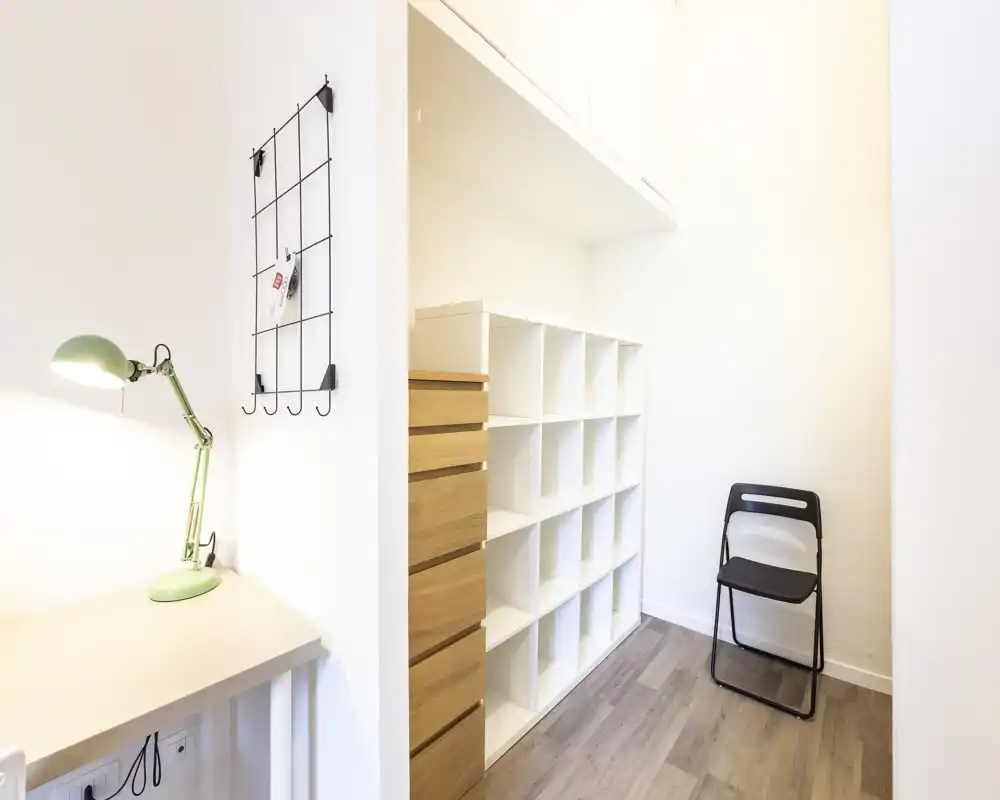 Pequeña entrada luminosa con estanterías empotradas, un escritorio compacto con lámpara verde y una silla plegable: espacio de llegada ordenado y funcional.