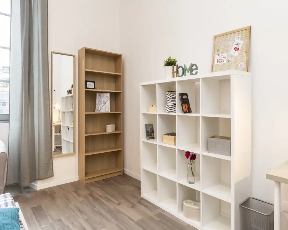 Área de estar luminosa y moderna con estantería de cubos, una librería alta de madera y un espejo de cuerpo entero; decoración neutra y luz natural que crean un ambiente limpio y acogedor.