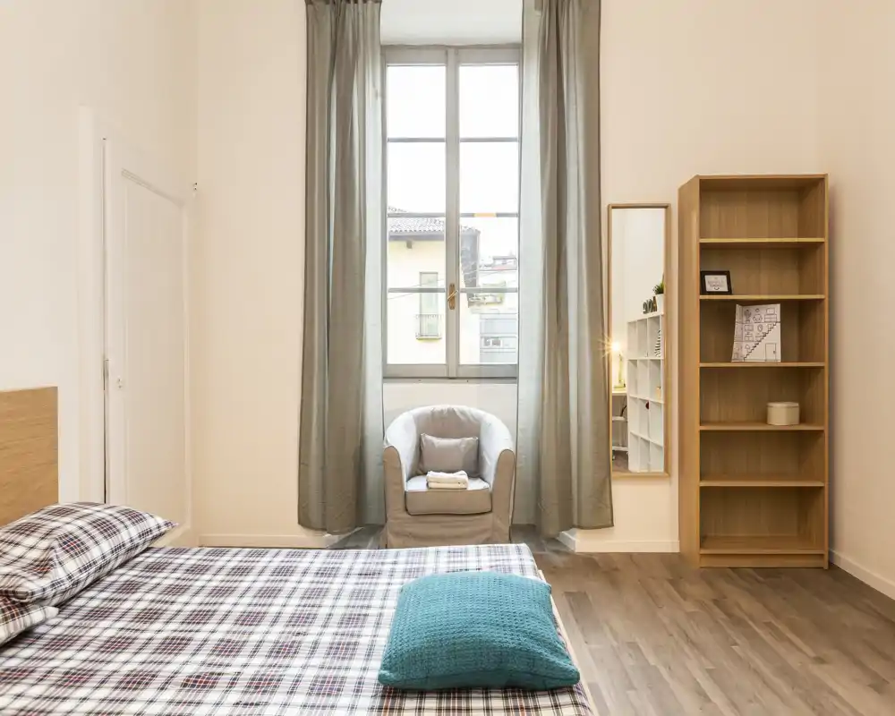 Dormitorio luminoso y bien decorado con una gran ventana central, un sillón cómodo y una cama arreglada con ropa a cuadros — ideal para una estancia urbana acogedora.