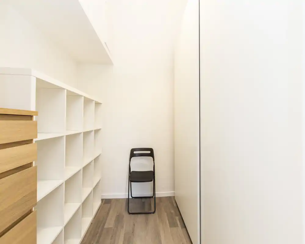 Un pequeño vestidor o nicho de almacenamiento con estanterías blancas integradas, una unidad de cajones de madera a la izquierda, un gran armario blanco a la derecha y una silla plegable contra la pared del fondo. Espacio limpio, minimalista y bien iluminado, ideal para almacenamiento organizado.