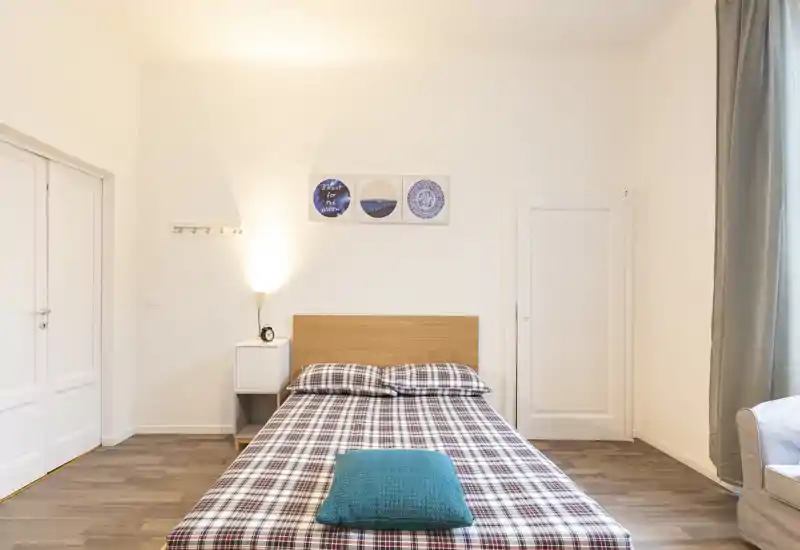 Dormitorio luminoso y minimalista con cama doble centrada, ropa de cama de cuadros y cojín turquesa; mesita sencilla y lámpara de pared que aportan calidez y estilo moderno.