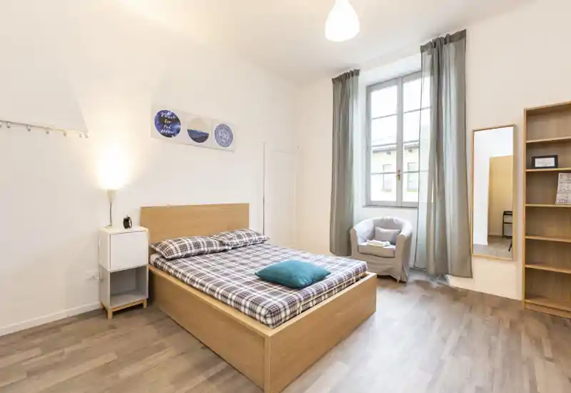 Dormitorio luminoso y ordenado con cama doble de madera, mesita de noche y un cómodo sillón junto a la ventana.