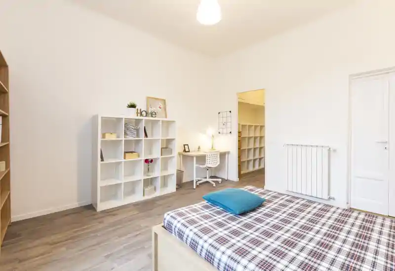 Dormitorio moderno y luminoso con cama bien hecha en primer plano, estantería y pequeño escritorio; decoración minimalista y mucha luz, perfecto para un anuncio de alquiler.