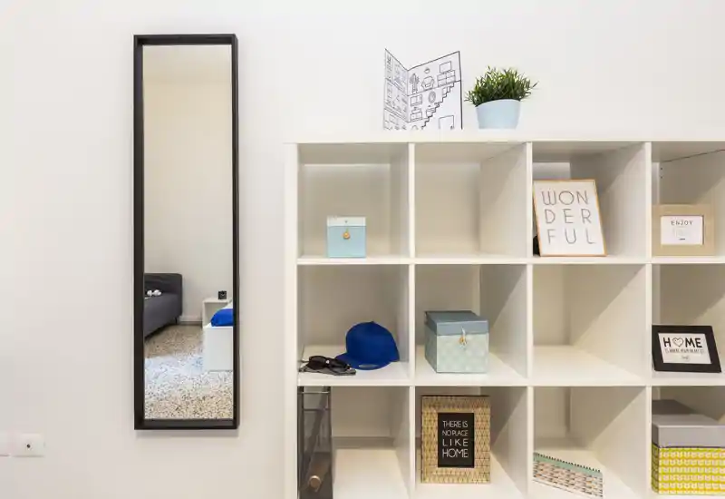 Coin de chambre soigné avec une étagère cubique et une grande glace reflétant le lit et la table de chevet ; quelques accessoires discrets pour un style moderne et épuré.