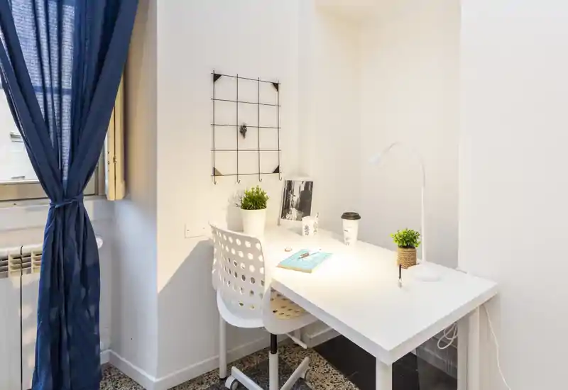 Coin bureau lumineux avec bureau blanc, chaise ergonomique, lampe de bureau et petites plantes près d'une fenêtre avec rideau — idéal pour travailler ou lire.