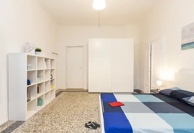Chambre moderne et lumineuse avec grand lit au premier plan, armoire blanche et étagère murale; style minimal et lampe de chevet chaleureuse.
