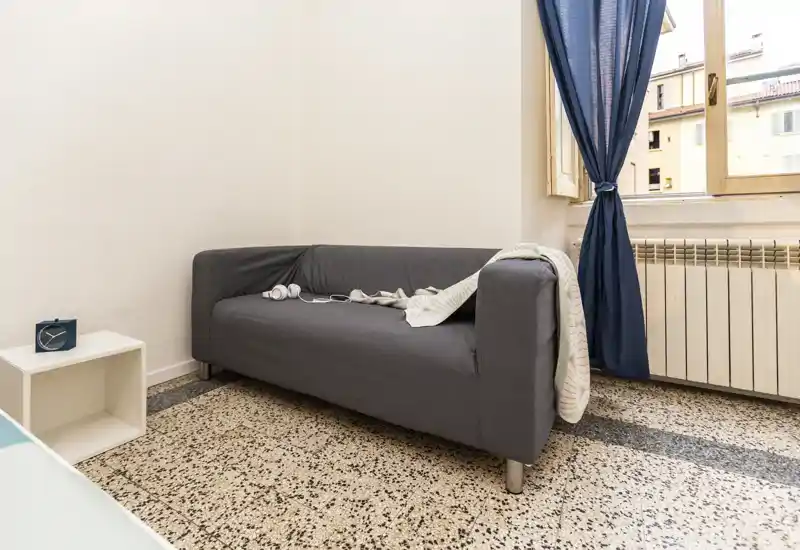 Coin salon compact avec un canapé gris deux places, lumière naturelle venant de la fenêtre et textiles simples — idéal pour un séjour urbain confortable.