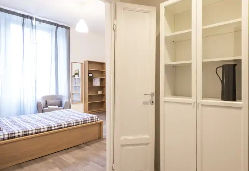 Chambre lumineuse et minimaliste avec lit en bois, fauteuil et étagère; grande fenêtre apportant une lumière naturelle douce et une ambiance moderne et nette.