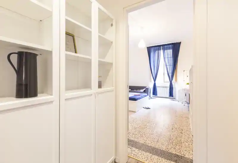 Vue depuis le couloir vers une chambre lumineuse aux rideaux bleus et un lit simple partiellement visible ; étagères blanches et sol en terrazzo clair offrent une ambiance propre et aérienne.