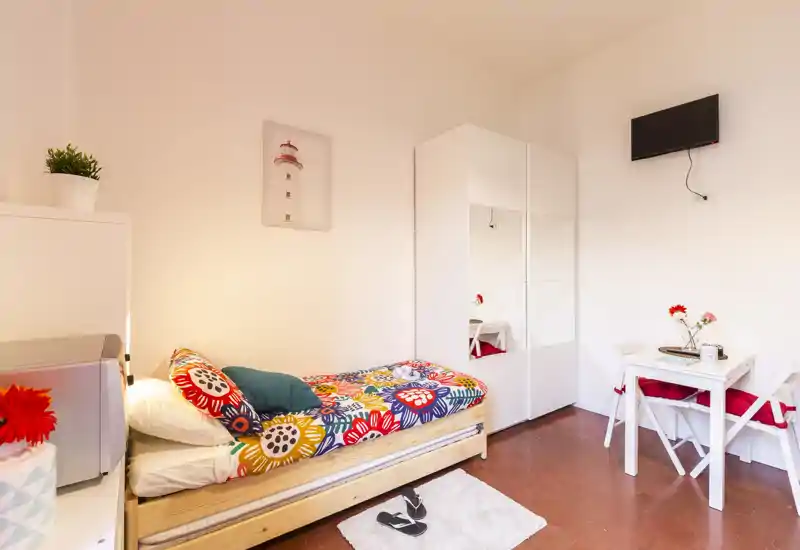 Camera singola luminosa e ben arredata con letto dai colori vivaci, armadio bianco con ante a specchio, piccolo tavolo e TV a parete — perfetta per un soggiorno accogliente.