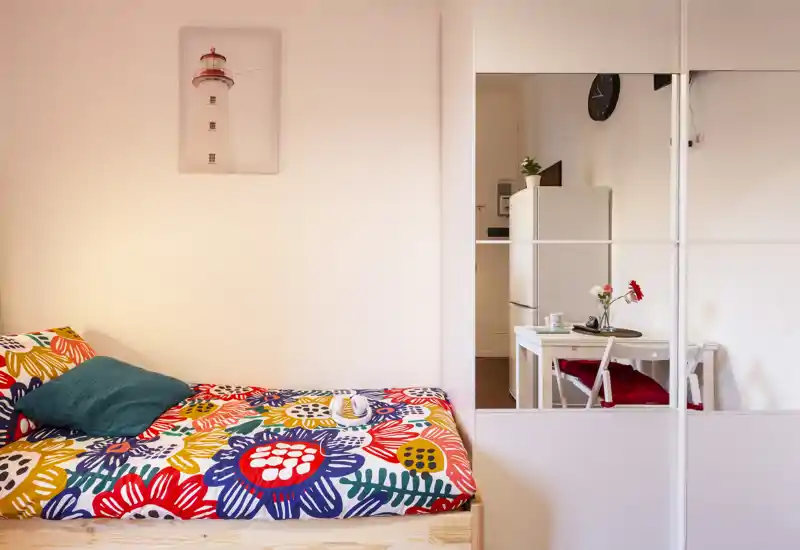 Camera da letto luminosa e colorata con un letto singolo con copriletto floreale vivace e cuscino decorativo; l'armadio con ante a specchio aggiunge profondità e riflette un piccolo angolo pranzo.
