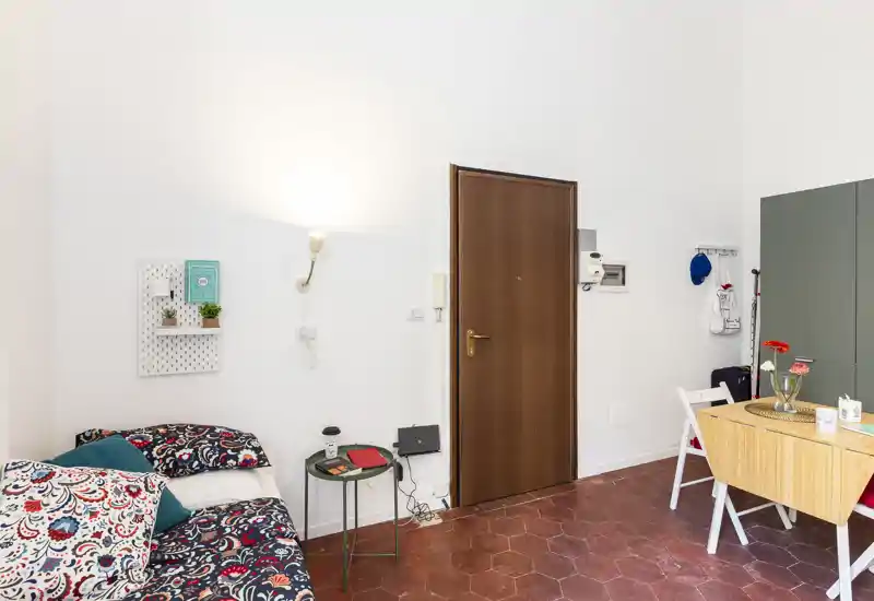 Zona de estar luminosa y ordenada con textiles estampados en un sofá cama, una mesita auxiliar y un pequeño comedor junto a la entrada — ideal para un acogedor apartamento en la ciudad.