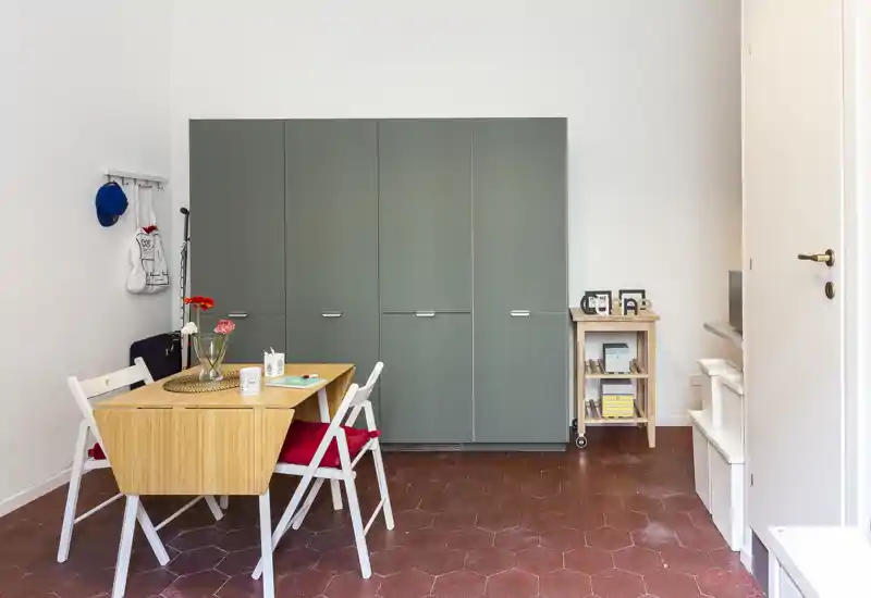 Piccolo ambiente living/pranzo con un tavolo pieghevole in legno, due sedie pieghevoli con cuscini rossi e un grande armadio grigio contro la parete bianca. La stanza è ordinata e luminosa con un arredamento semplice d'ispirazione scandinava.
