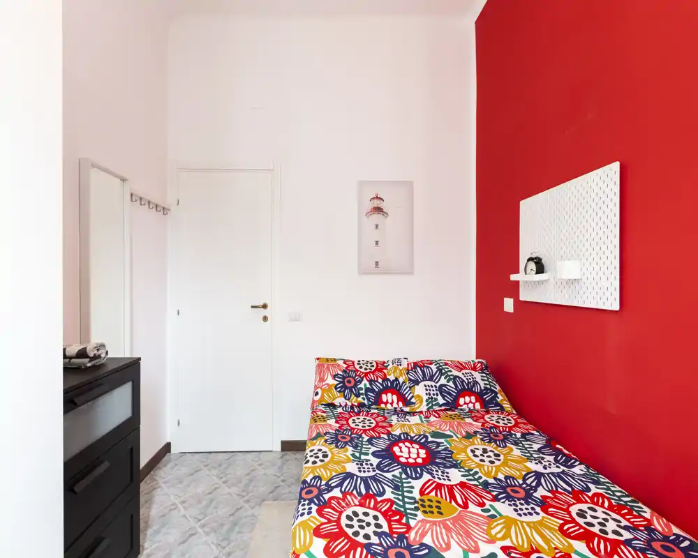 Bright, colorful bedroom with a patterned bedspread against a red accent wall; compact layout with a dresser and mirror, ideal for single occupancy or short stays.