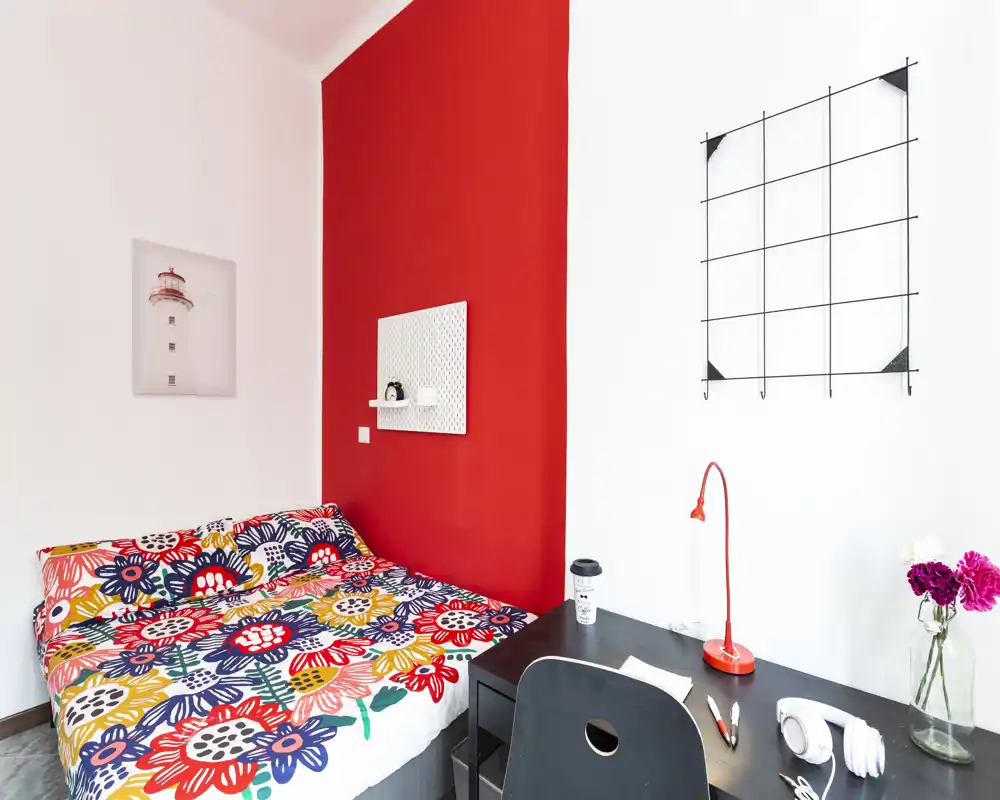 Bright, colorful bedroom with a double bed dressed in a bold floral duvet, a small workspace with desk and lamp, and decorative wall accents creating a cheerful, modern feel.