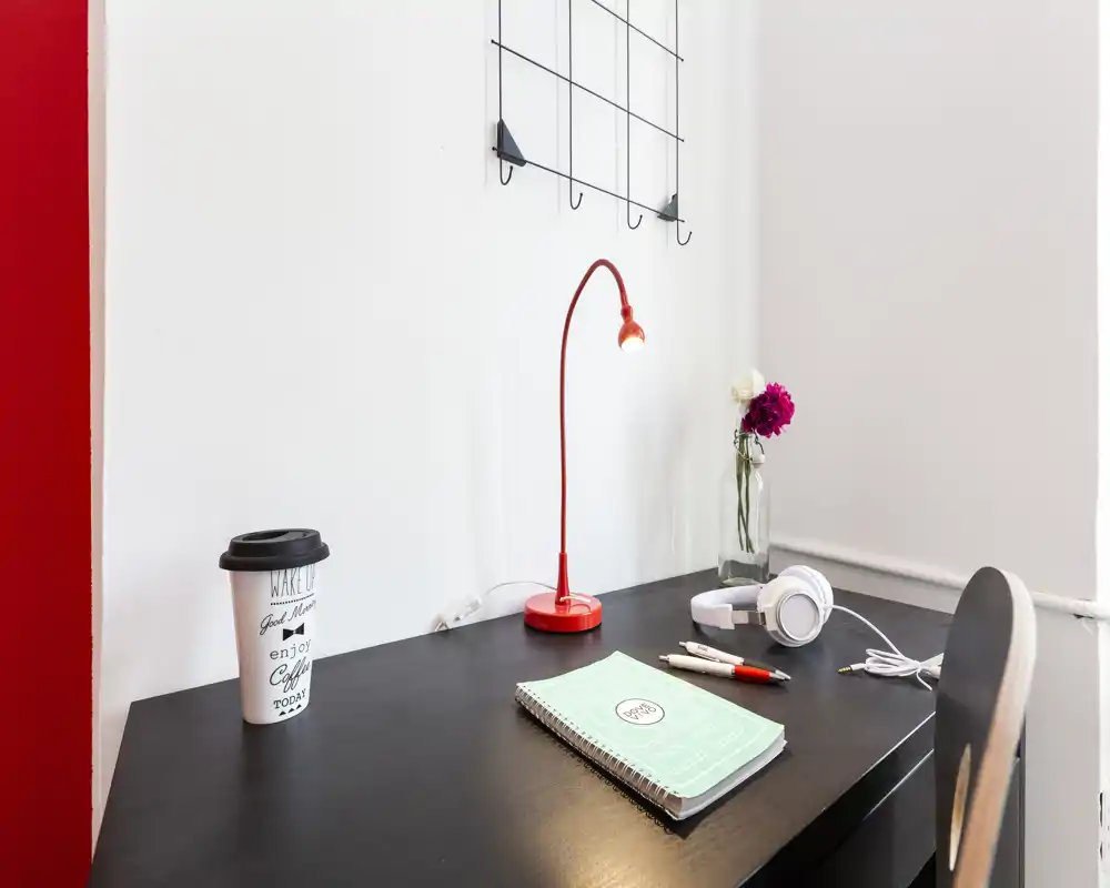 Compact workspace corner with a dark desk, red task lamp, headphones and a notebook — bright, tidy and styled for short-term rentals or study.