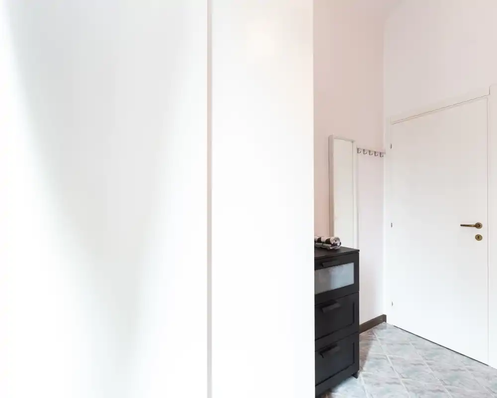 Bright, minimal entrance with a white door, coat hooks, a tall mirror and a small black chest of drawers on tiled flooring—clean modern entryway.