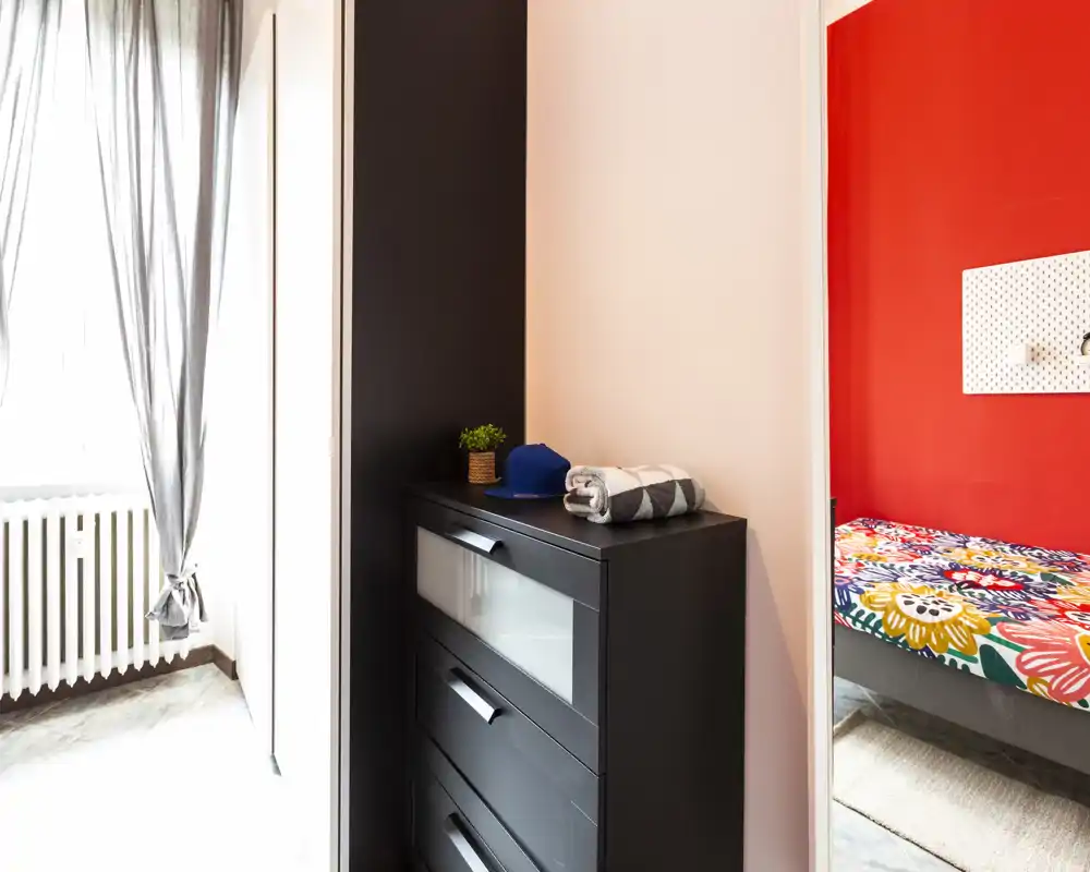 Bright, modern bedroom corner with a colorful patterned bed and a black chest of drawers beside a window letting in natural light.