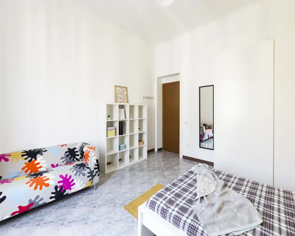 Camera da letto luminosa e moderna con un divano colorato, scaffalatura bianca e un letto matrimoniale con copriletto a quadretti; spazio minimale e ben illuminato, ideale per annunci.