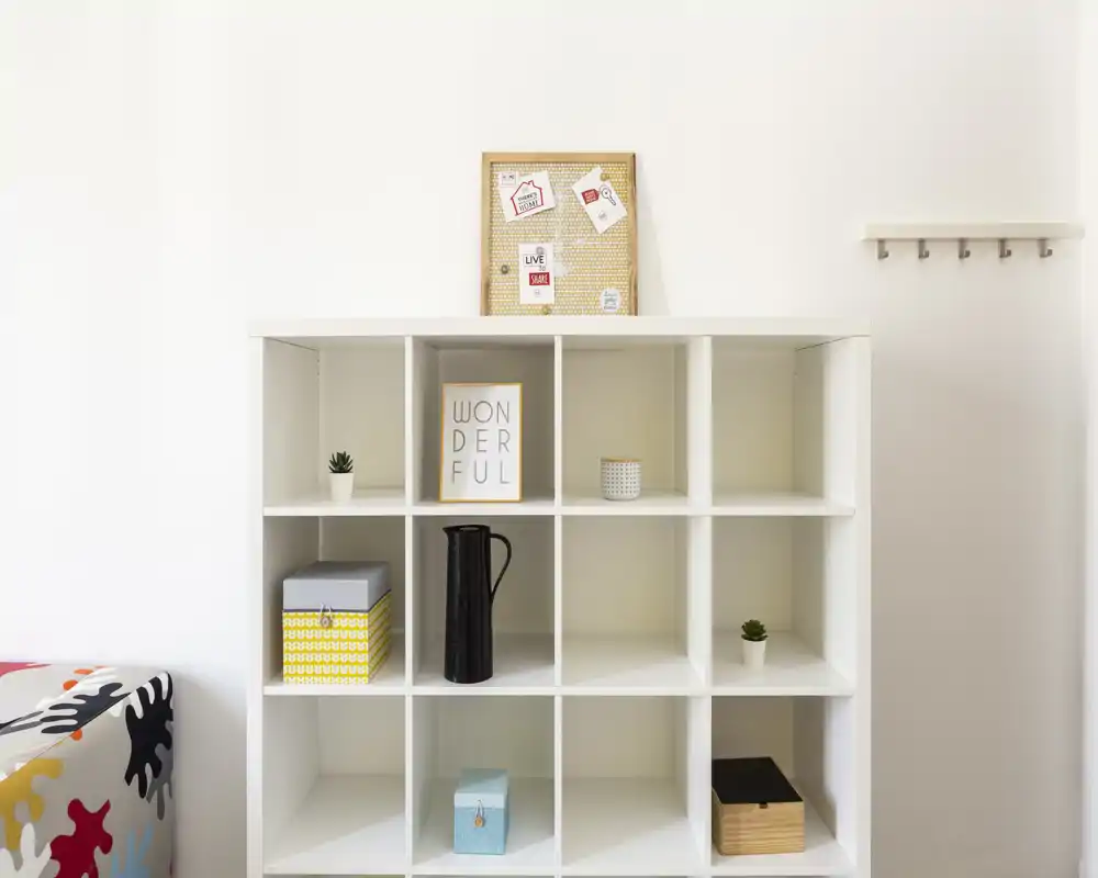 Zona giorno luminosa e minimal con una libreria a cubi bianca, piccoli oggetti decorativi e un accenno di divano colorato — stile moderno e pulito adatto per annunci di appartamento.
