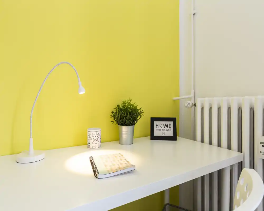 Angolo scrivania luminoso e minimale con lampada bianca, quaderno, piccolo vaso e cornice su una parete gialla: perfetto per un compatto spazio di lavoro domestico.