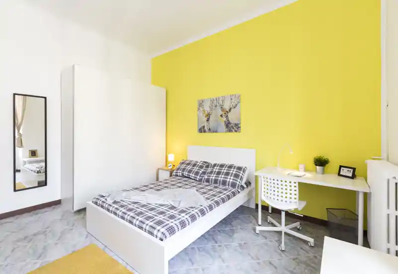 Camera da letto luminosa e moderna con letto matrimoniale, biancheria a quadri, scrivania bianca e armadio su una vivace parete gialla — perfetta per un affitto accogliente.