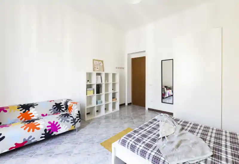 Camera da letto luminosa e moderna con un divano colorato, scaffalatura bianca e un letto matrimoniale con copriletto a quadretti; spazio minimale e ben illuminato, ideale per annunci.