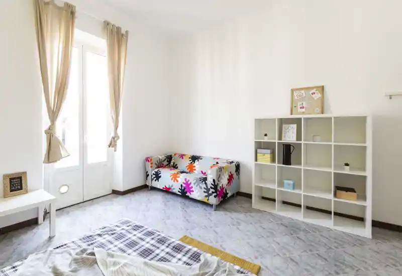 Camera da letto luminosa e minimal con un divano a due posti colorato, scaffale modulare e letto parzialmente visibile in primo piano; ampia finestra con tende beige che garantisce molta luce naturale.