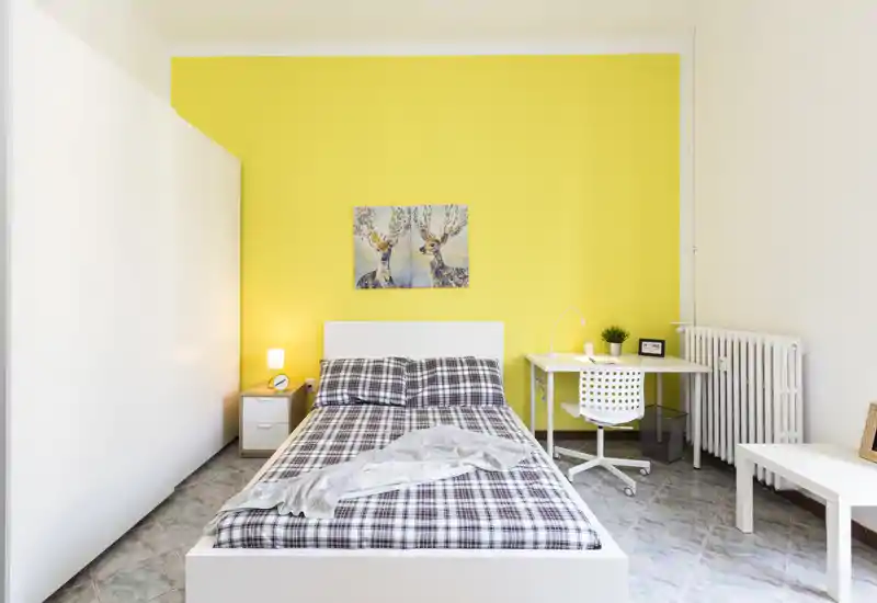 Camera da letto luminosa e moderna con letto matrimoniale a fantasia contro una parete gialla vivace, comodino con lampada e una scrivania compatta con sedia — perfetta per viaggiatori singoli o coppie che cercano una stanza ordinata e ben illuminata.