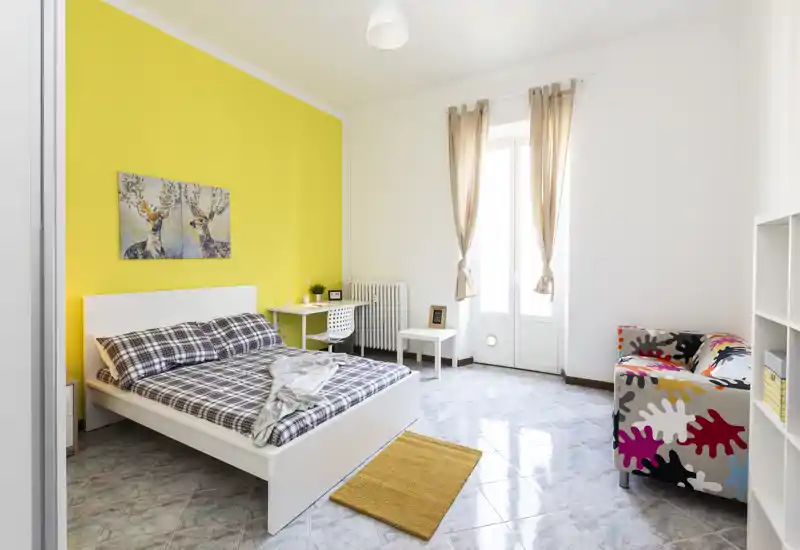 Camera da letto luminosa e di design con struttura del letto bianca su una parete gialla, biancheria a quadri, una scrivania e una poltrona colorata — perfetta per annunci in affitto che puntano su luce e stile.