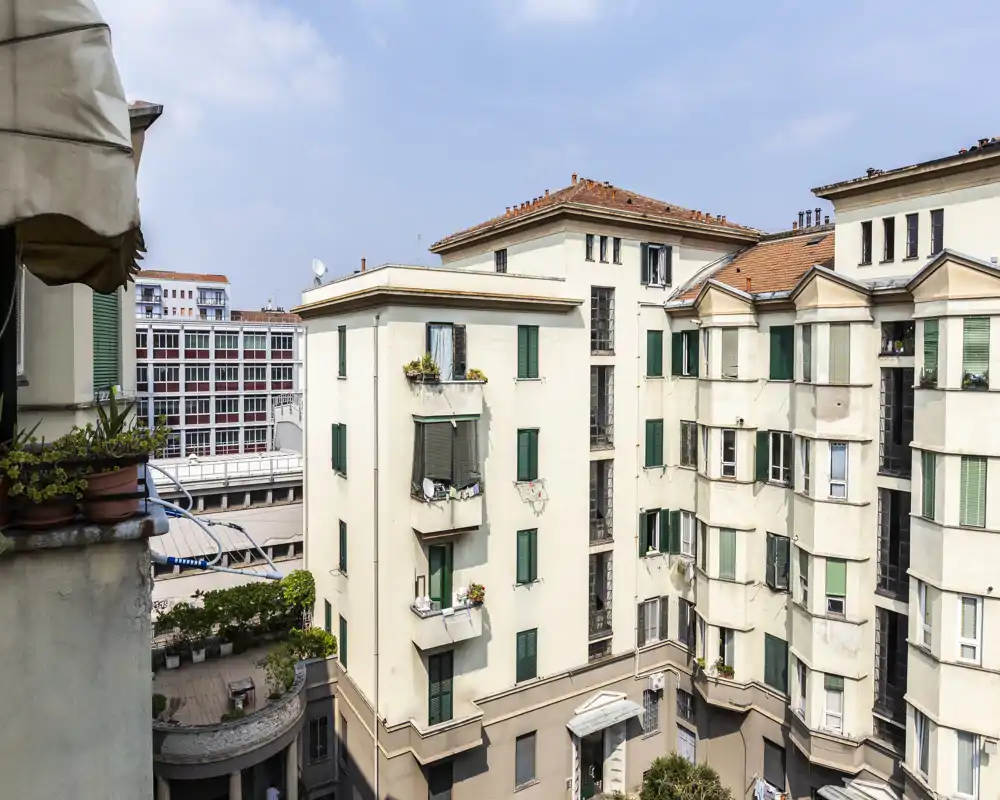 Exterior courtyard and neighboring apartment façades seen from a balcony; potted plants and a partial railing visible in the foreground with clear daylight.