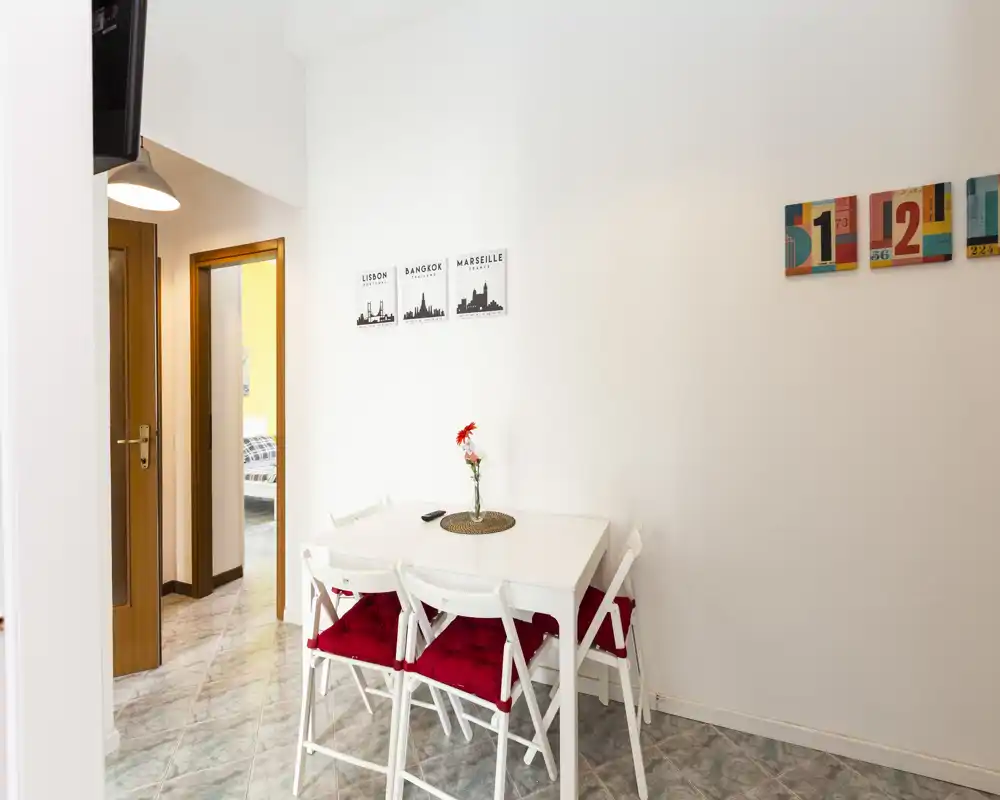 Compact dining nook with a white table and four chairs accented by red cushions and a simple vase centerpiece. Clean, bright walls and minimalist decor create a welcoming spot for meals.