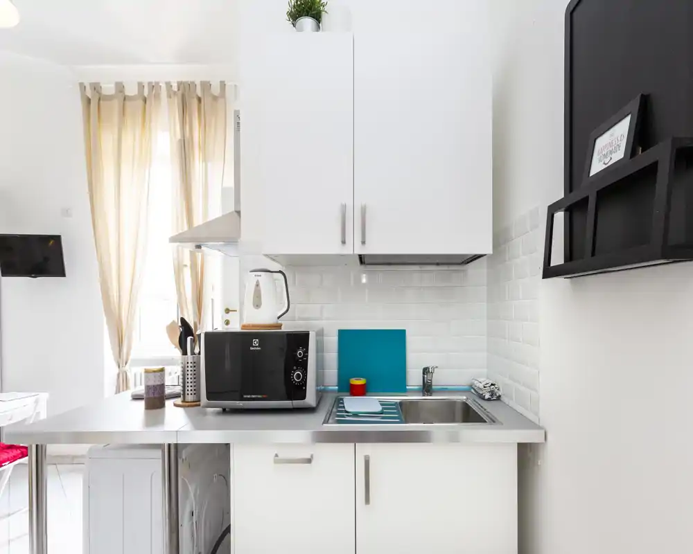 Kitchenette moderna e compatta con pensili bianchi, lavello in acciaio, microonde e una finestra luminosa che lascia entrare luce naturale; ordinata e ben attrezzata per soggiorni brevi.