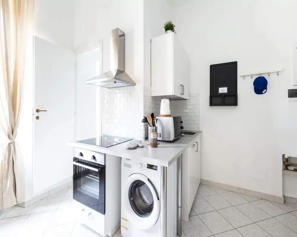 Cucina moderna e compatta, luminosa, con forno, piano a induzione, cappa e lavatrice integrata; pensili bianchi e rivestimento a piastrelle per un aspetto pulito.