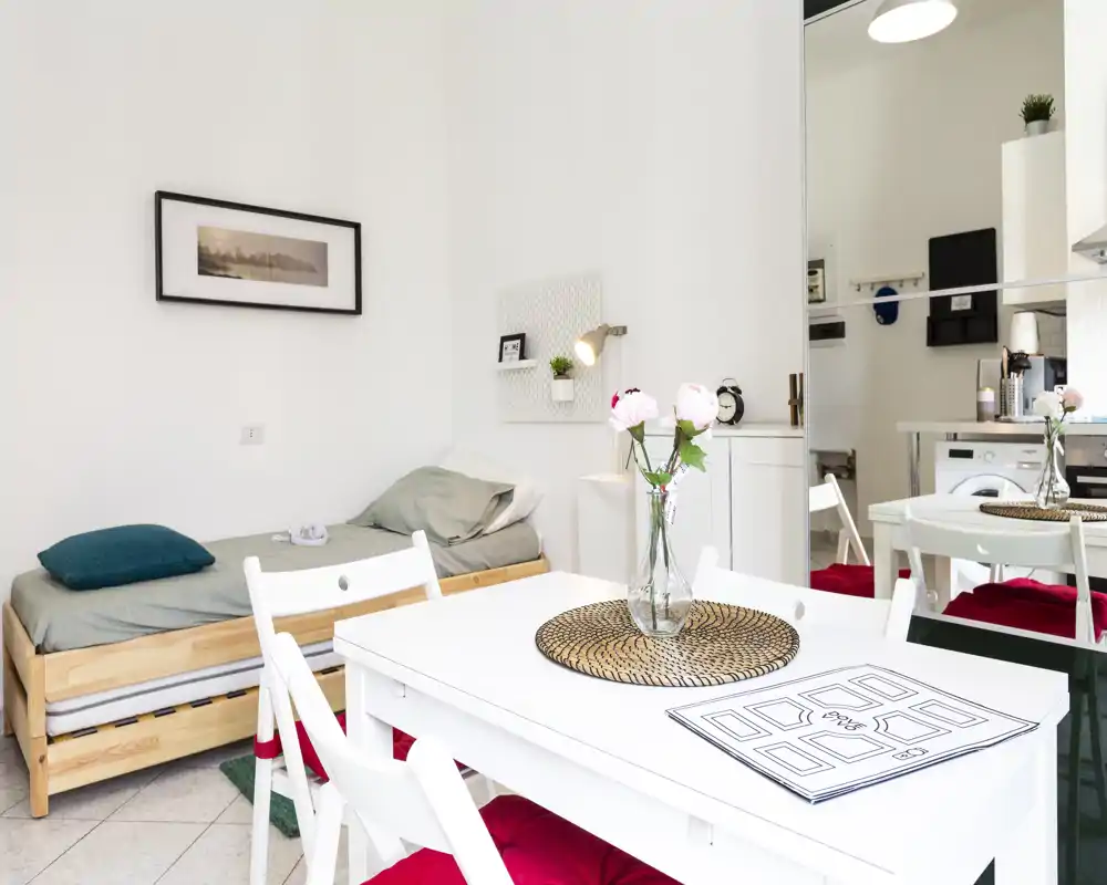 Camera da letto/studio luminosa e minimal con letto singolo in legno, tavolo da pranzo bianco e sedie con cuscini rossi; fiori freschi sul tavolo per un tocco accogliente.