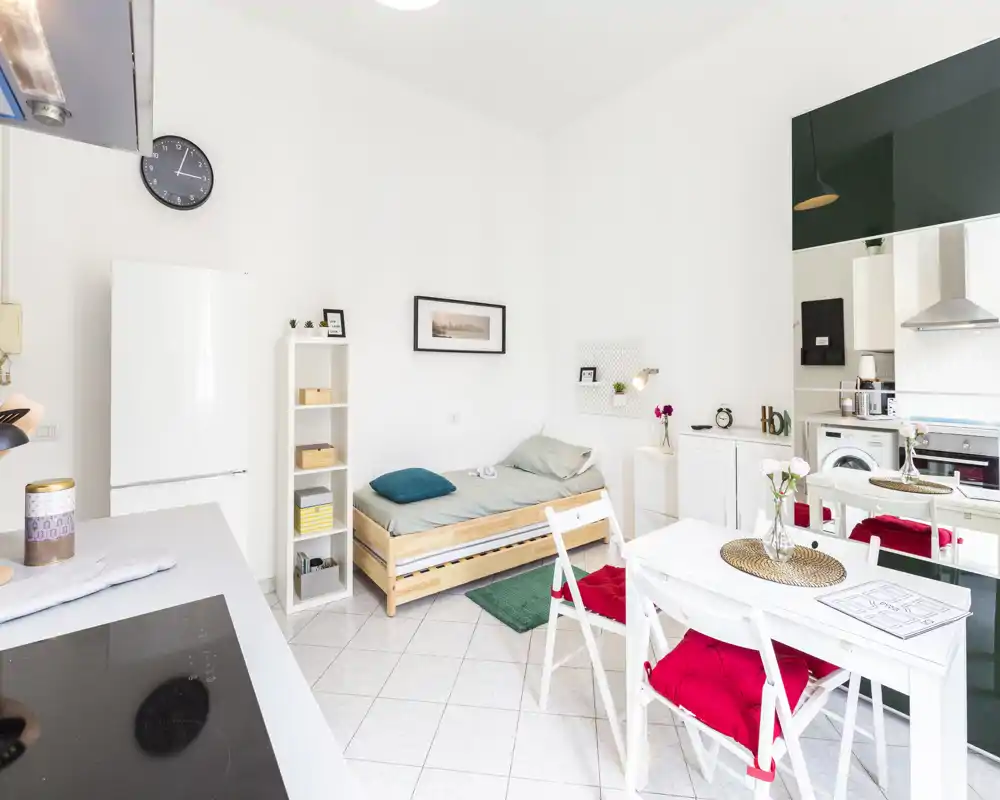 Monolocale luminoso con letto singolo a parete, angolo cottura compatto e piccolo tavolo da pranzo. Arredamento bianco e pulito con tocchi di colore su cuscini e tappeto.