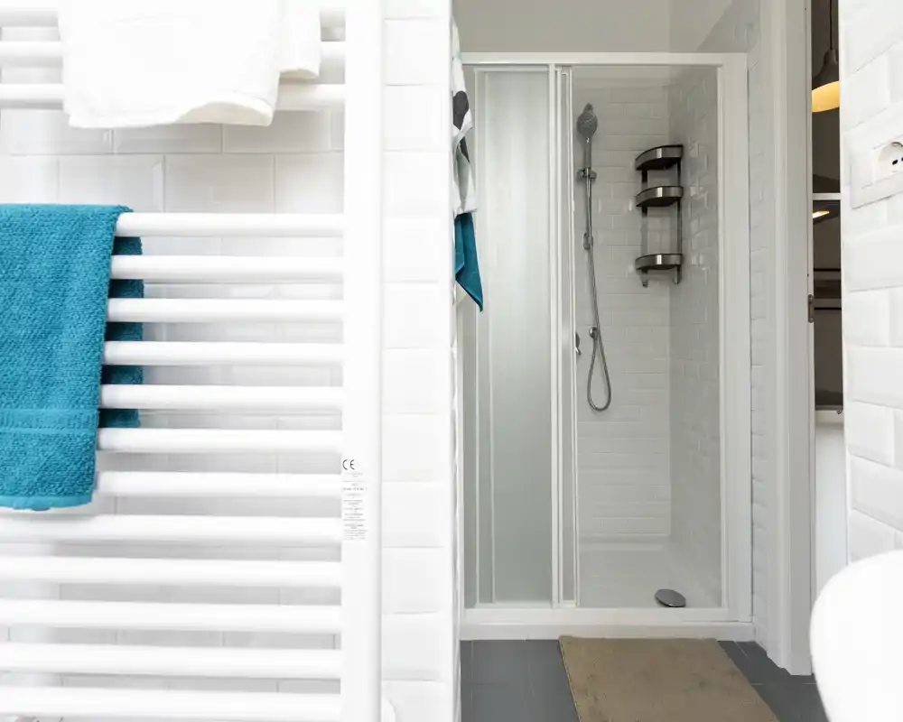 Bagno moderno e luminoso con cabina doccia piastrellata, porta scorrevole in vetro, doccia a mano e mensole a muro. Piastrelle bianche e scaldasalviette con asciugamani turchesi per un aspetto pulito e fresco.