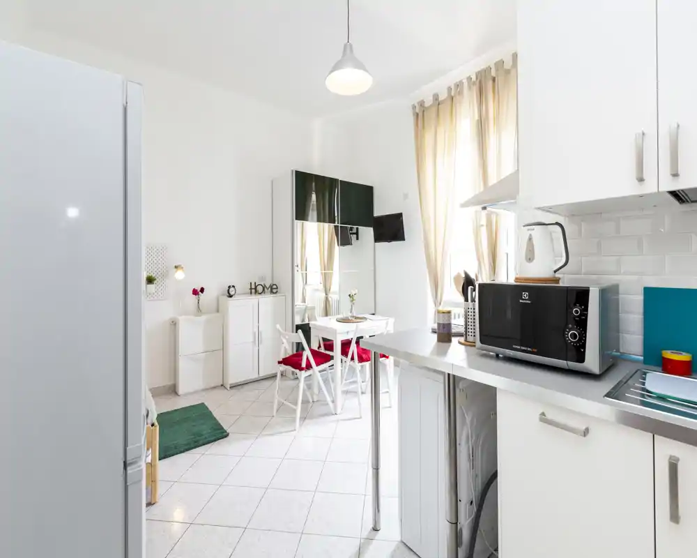 Cucina moderna e luminosa in open space con piccolo tavolo da pranzo, microonde sul piano e luce naturale proveniente da alte finestre con tende leggere.