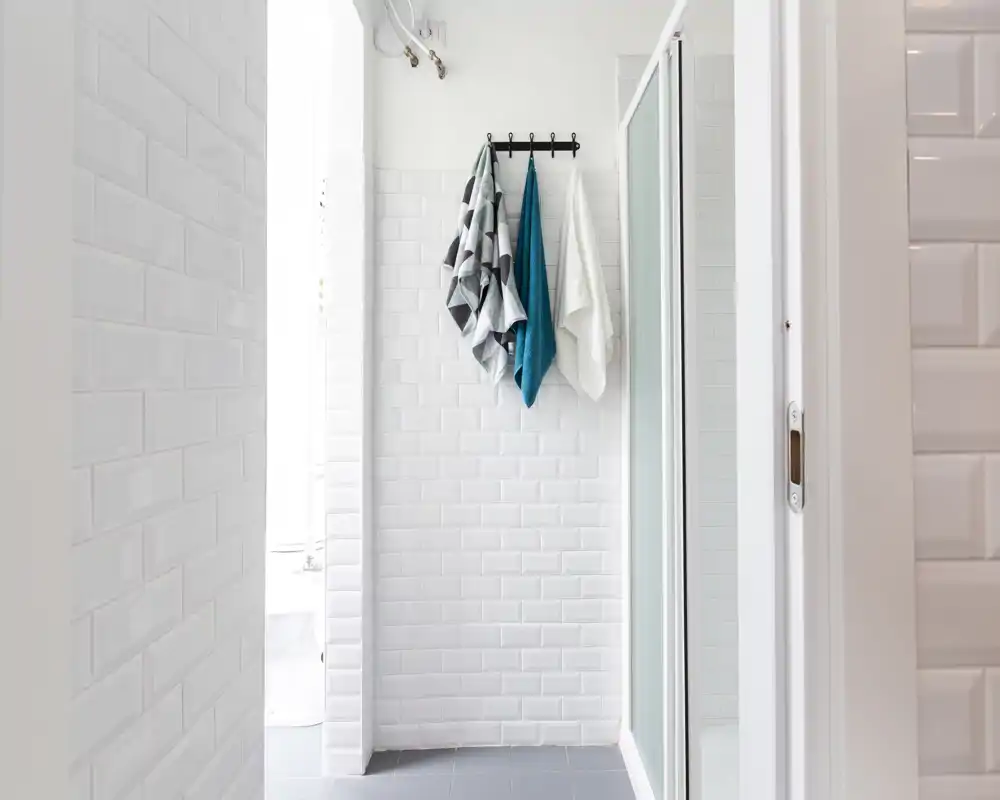 Bagno moderno e pulito con piastrelle bianche a sottosquadra, porta doccia in vetro e tre asciugamani appesi a un attaccapanni a parete.