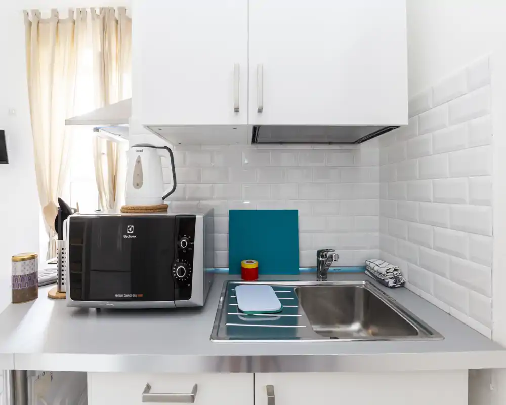 Angolo cottura moderno e compatto con lavello in acciaio, forno a microonde sul piano e rivestimento a mattonelle bianche — luminoso e ordinato, ideale per soggiorni brevi.