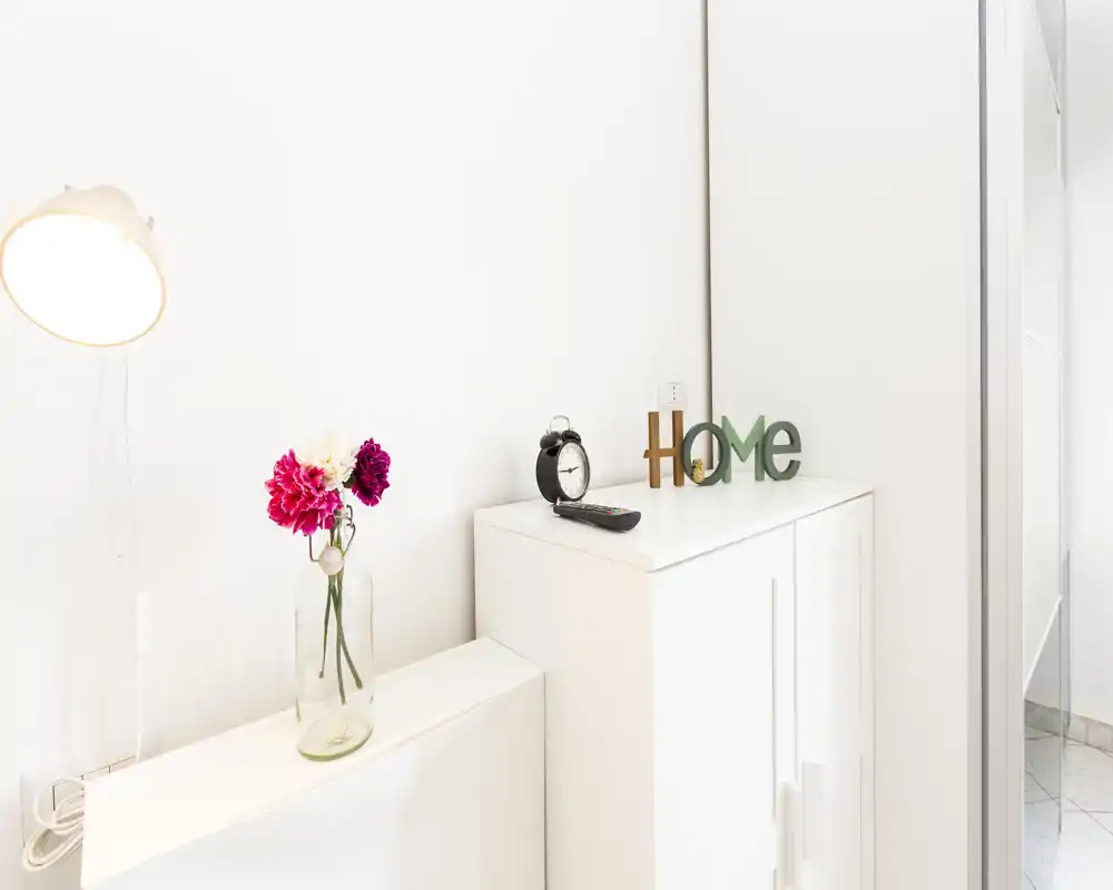 Ingresso luminoso e minimale con un mobiletto bianco, lettere decorative "Home", una sveglia e un vaso di fiori — un angolo d'ingresso pulito e accogliente.