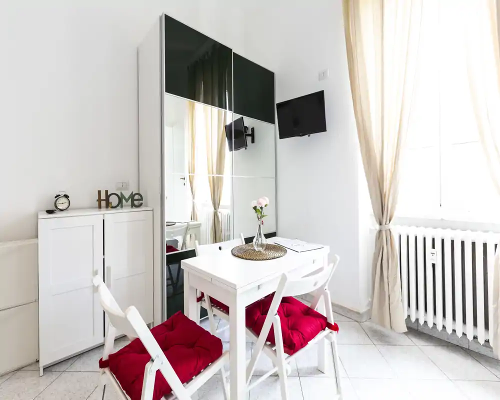 Accogliente angolo pranzo luminoso con tavolo bianco e due sedie con cuscini rossi, tende calde e un armadio specchiato che amplia la stanza.