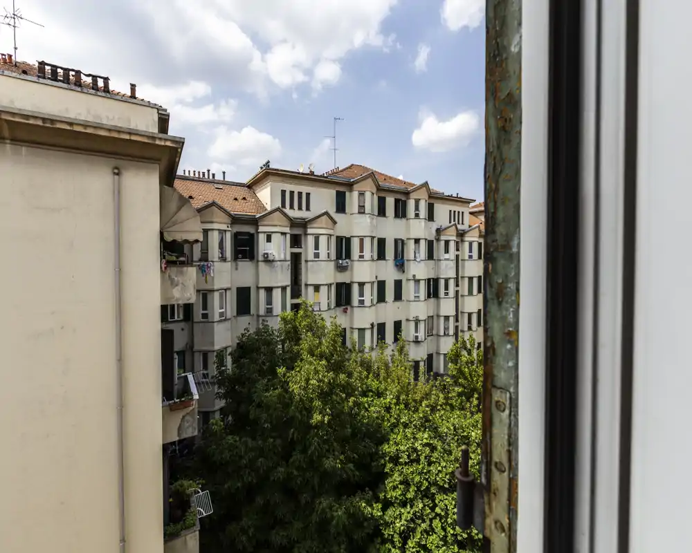 Vista cittadina dalla finestra con palazzi residenziali vicini, alberi rigogliosi in primo piano e un cielo parzialmente nuvoloso luminoso.